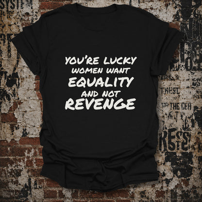 A black short-sleeve graphic T-shirt laid flat against a distressed brick and concrete wall. The shirt has bold white text reading, 'YOU'RE LUCKY WOMEN WANT EQUALITY AND NOT REVENGE.