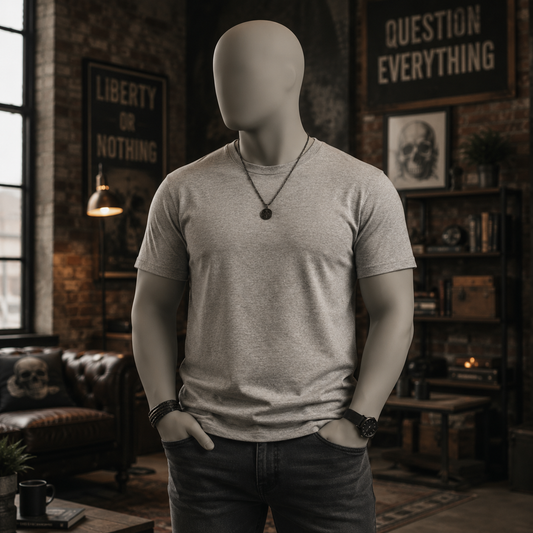 A light gray heather crewneck t-shirt displayed on a headless mannequin torso in a dark, rustic loft-style room with brick walls, posters, and shelves.